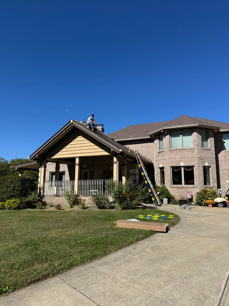 Large brick home in daylight with a ladder at the porch and an installer up on the roof