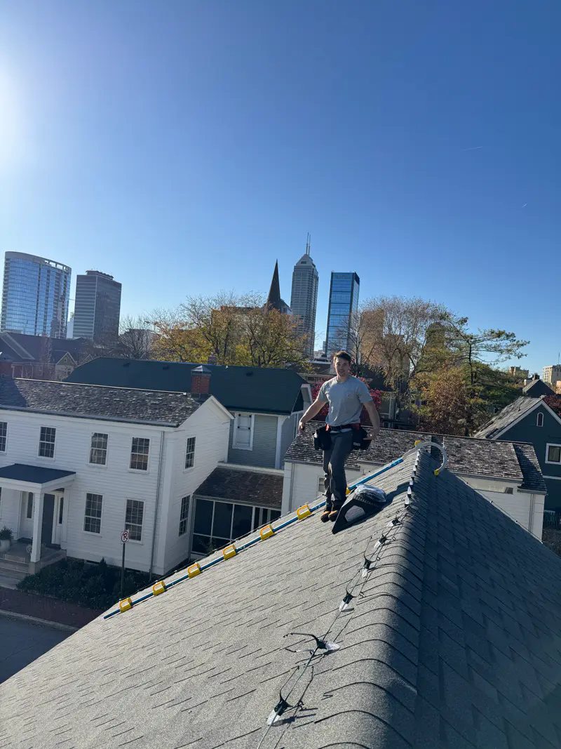 Kyle standing on a shingled roof with a ladder hooked over the ridge and the downtown Indianapolis skyline behind him